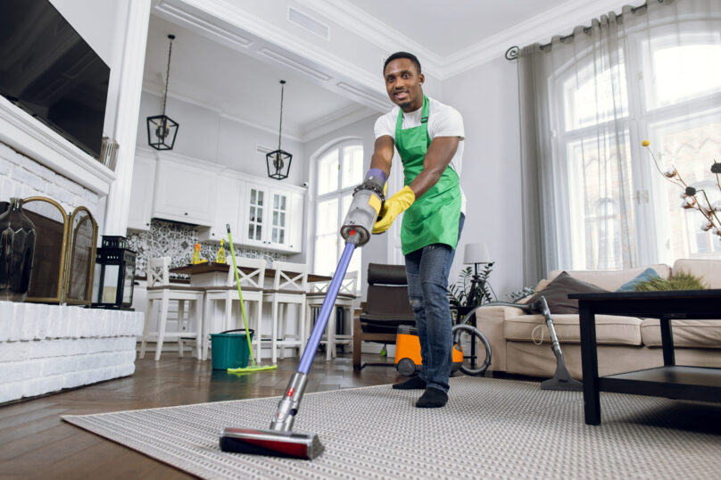 African man removing dust from soft carpet with modern vacuum cleaner. Male cleaner wearing green apron and yellow rubber gloves. Working process at bright room.